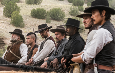 A line of men wearing cowboy hats and period clothing riding horses through a hilly landscape.