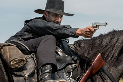 A man in a black hat and shirt aims a silver revolver while mounted on a black horse.
