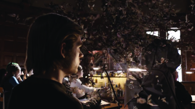 A man in profile looks at a large, dark, textured installation in a dimly lit indoor space.