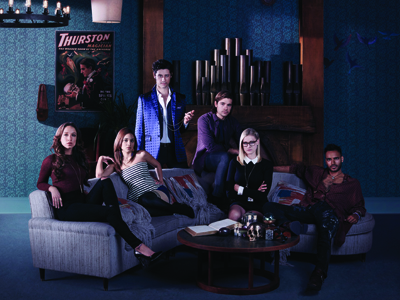 Six young adults pose in a dark room with a blue patterned wall and a wooden mantelpiece.
