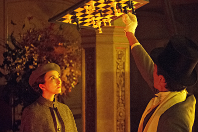 A man in a top hat and white gloves holds a chessboard aloft while a woman in a grey beret watches him.