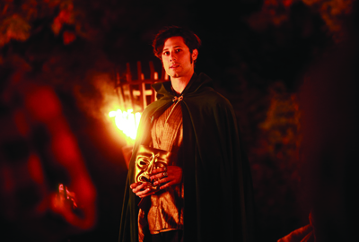 A man in a dark green cloak holds a gold mask while standing near a bright orange flame in the dark.