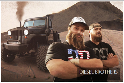 Two bearded men in black t-shirts stand before a large, black custom off-road vehicle in a desert landscape.