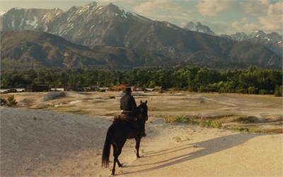 A person on a dark horse rides across a sandy plain toward a lush forest and large mountains.