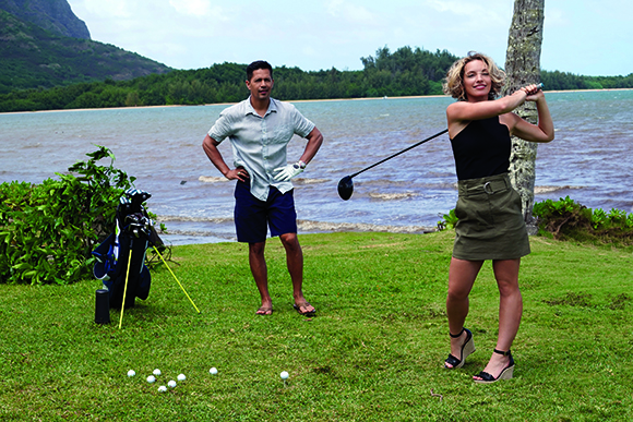 A woman swings a golf club on a lawn near a beach while a man stands behind her watching.