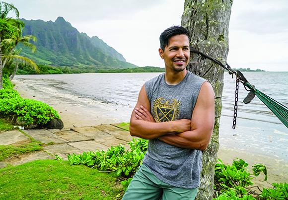 A smiling man in a grey sleeveless shirt leans against a palm tree on a tropical beach with mountains in the background.