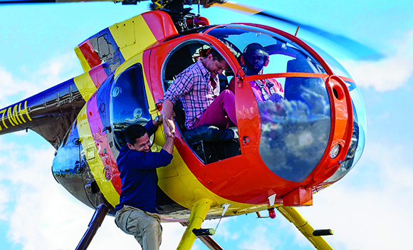 A man in a blue shirt helps a passenger in a plaid shirt board a bright orange and yellow helicopter.
