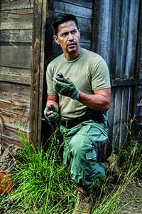 A man in a green t-shirt and cargo pants crouches near a wooden building, holding a small object.