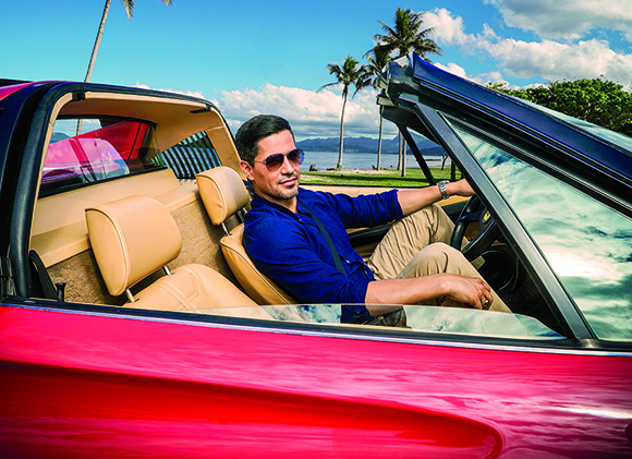 A man in a blue shirt and sunglasses sits in the driver's seat of a red convertible.