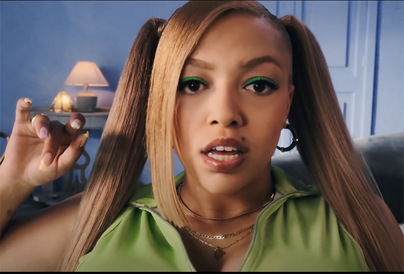 A woman with long blonde pigtails, green eyeliner, and a green top looks toward the camera.