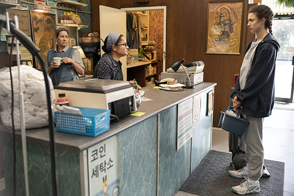 Two women behind a service counter talk to a young woman standing in a laundromat.