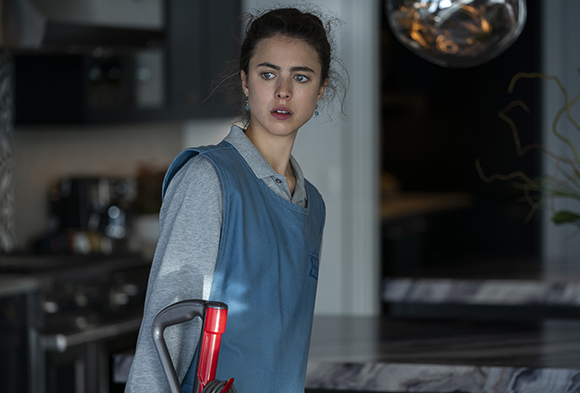 A young woman wearing a blue apron and grey shirt looks sideways with a worried expression in a kitchen.