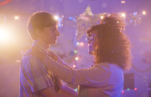A young man and woman stand close together in a room filled with soft purple light and festive decorations.