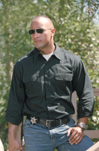 A man wearing sunglasses, a black button-down shirt, and blue jeans standing in front of green foliage.