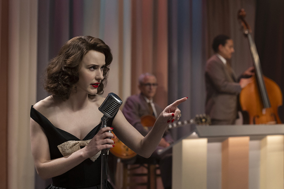 A woman in a black dress sings into a vintage microphone on a stage with musicians in the background.