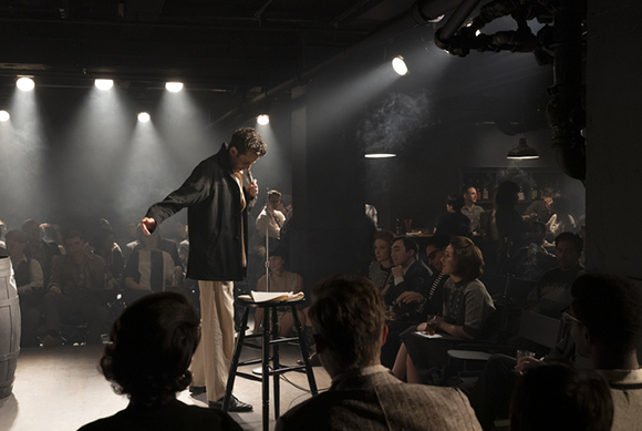 A man in a black jacket stands at a microphone on a small stage in a dark, smoky comedy club.