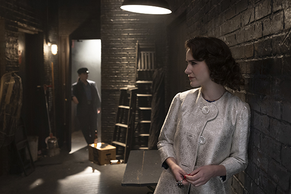 A woman in a white jacket leans against a dark brick wall in a dimly lit corridor.