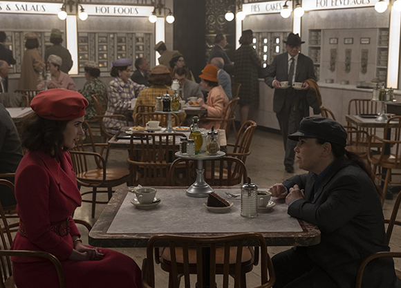 A woman in a red coat and beret sits across from a woman in a dark suit at a diner table.