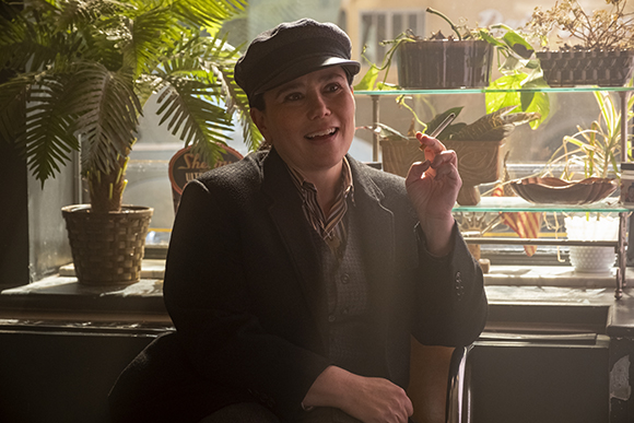 A person wearing a dark cap and jacket sits near plants, holding a lit cigarette.