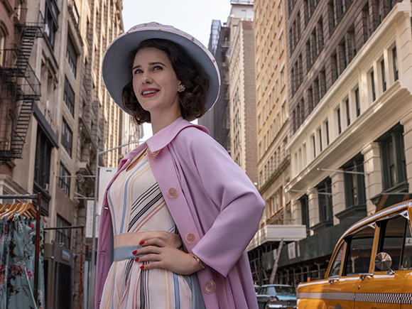A woman wearing a lavender hat and pink coat stands on a city street lined with tall buildings.