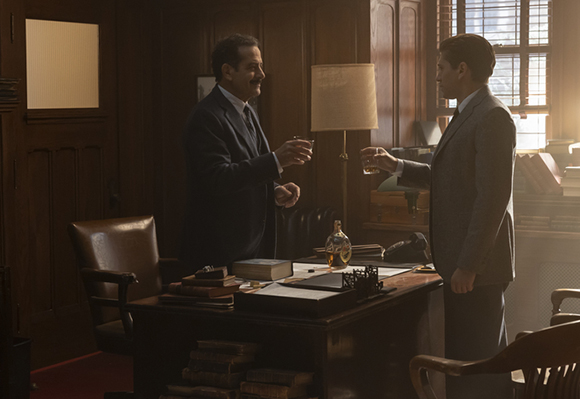 Two men in suits stand in a wood-paneled office, each holding a small glass.