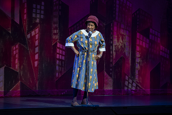 A woman in a patterned blue dress and red hat stands at a microphone on a stage with a city skyline backdrop.