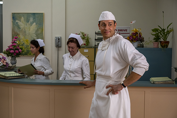 A man in a white uniform and cap stands in front of a counter while two women in white uniforms stand behind it.