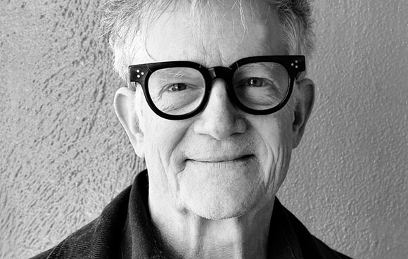 Black and white close-up portrait of an older man wearing thick-rimmed black glasses and smiling slightly.