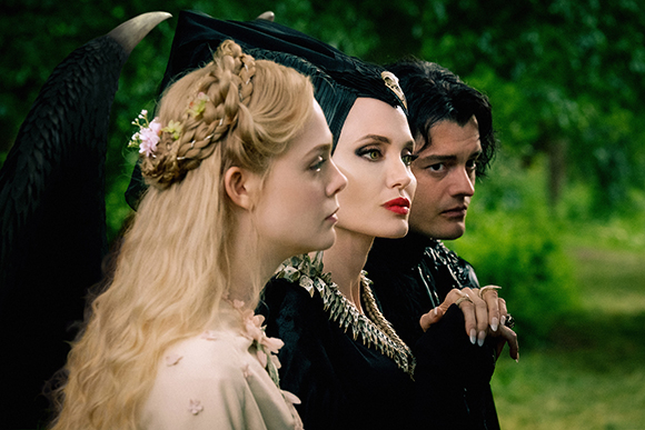A blonde woman in white, a woman in black with a headpiece, and a man in black look off-camera in a forest.