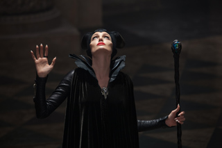 A woman in black horns and a dark gown looks up while holding a staff.