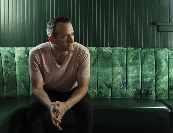 A man in a pink t-shirt sits on a green patterned booth against a dark green paneled wall.