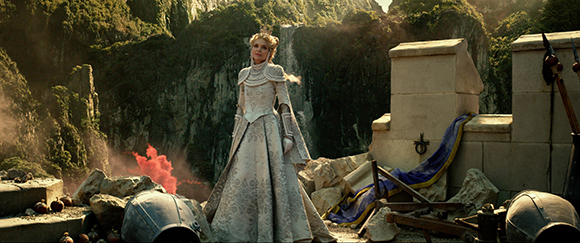 A woman in a white, patterned gown stands in a landscape of stone ruins and steep cliffs.