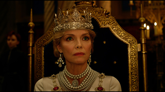 A woman wearing a silver crown and heavy pearl necklaces sits on a gold ornate throne.