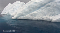 A massive white iceberg with jagged peaks floating in dark blue ocean water under a gray sky.