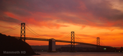 Silhouette of a large suspension bridge against a dramatic orange and purple sunset sky.