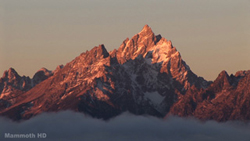 A sharp, snow-dusted mountain peak illuminated by orange sunset light above a layer of clouds.