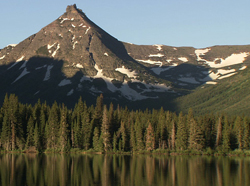 A sharp mountain peak with patches of snow rises above a dense evergreen forest and a calm lake.
