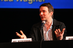 A man in a black jacket and striped shirt gestures with both hands while speaking into a microphone.
