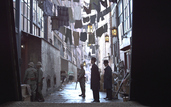 Men in period clothing stand in a narrow, cobblestone alleyway under hanging laundry lines.