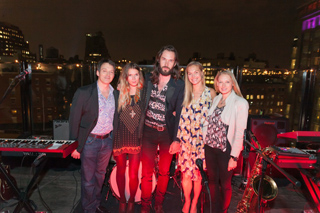 Five people stand together on a rooftop at night with musical instruments and city lights in the background.
