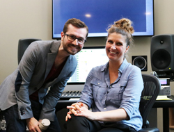 A man in a grey blazer and a woman in a blue shirt smile while sitting in a studio with monitors and speakers.