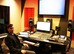 A man sits in a chair facing a large audio mixing desk equipped with monitors and computer screens.