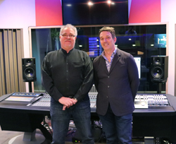Two men smiling while standing behind a large audio mixing console in a recording studio.