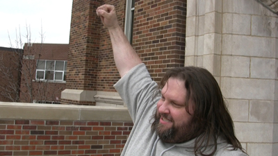 A man with long hair and a beard raises a clenched fist in the air outdoors.