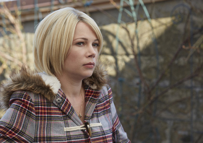 A blonde woman wearing a plaid coat with a fur-lined hood looks thoughtfully into the distance.