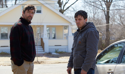 Two men stand in a residential area near a car and a yellow house.