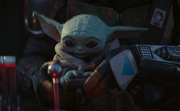 A small green creature with large ears being held by a person in dark armor.