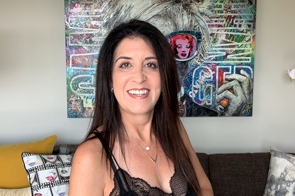 A woman with long dark hair and a black lace top smiles in front of a large, colorful abstract painting.