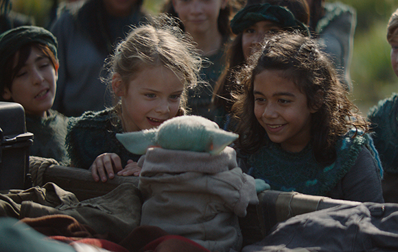 Three young children lean over a wooden cart to look at a small, green, long-eared creature.