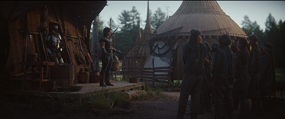 A woman in dark attire stands on a wooden platform addressing a group of people near large thatched huts.